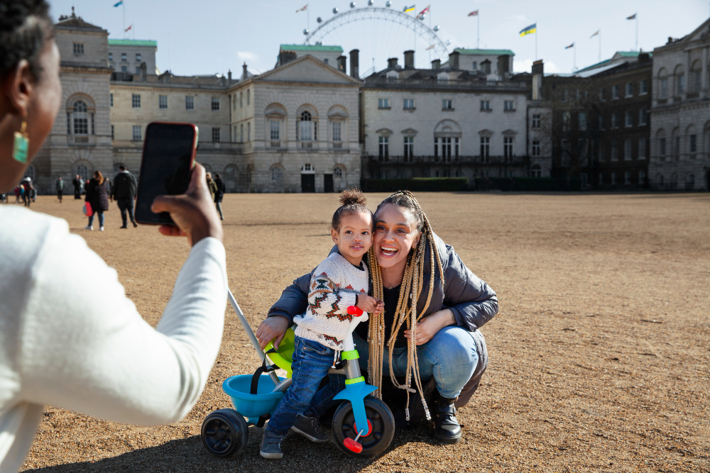 A woman captures a photo of a smiling child riding a tricycle in a sunny outdoor setting