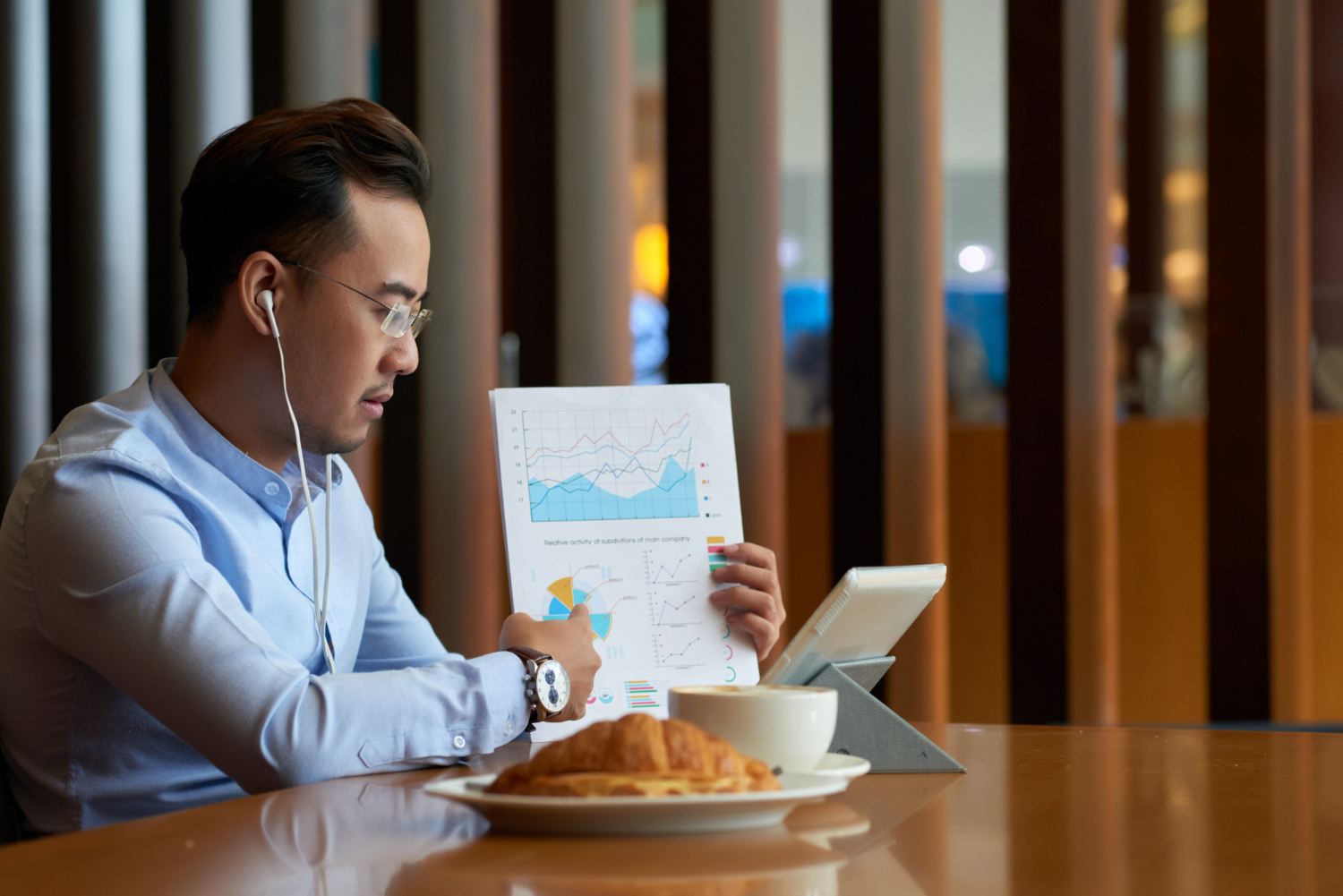 A man wearing a shirt and headphones is focused on reading a newspaper