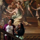 Two people admire a large classical painting while ascending a grand staircase