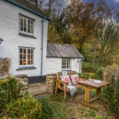 A cozy white cottage with a blue door, nestled in greenery, featuring an outdoor seating area with a table and cushions