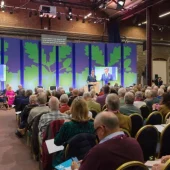 An audience watches a speaker on stage with a tree-themed backdrop at an indoor conference