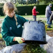A gardener in a green jacket empties a bucket of plant clippings, with others working in the background on a well-kept garden